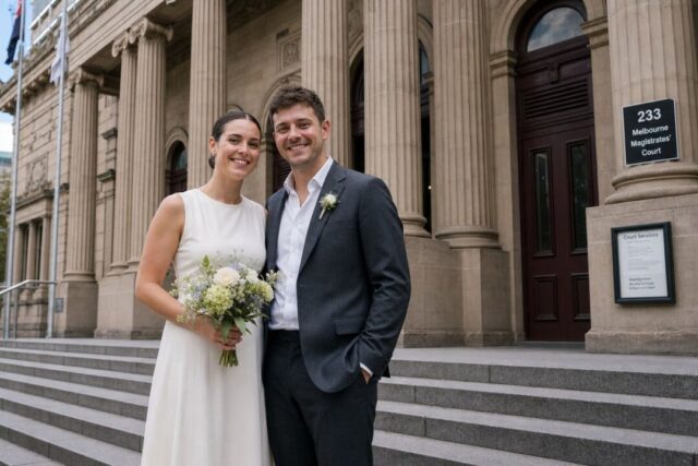 A couple in smart-casual wedding attire smiling on courthouse steps, from the knees up.