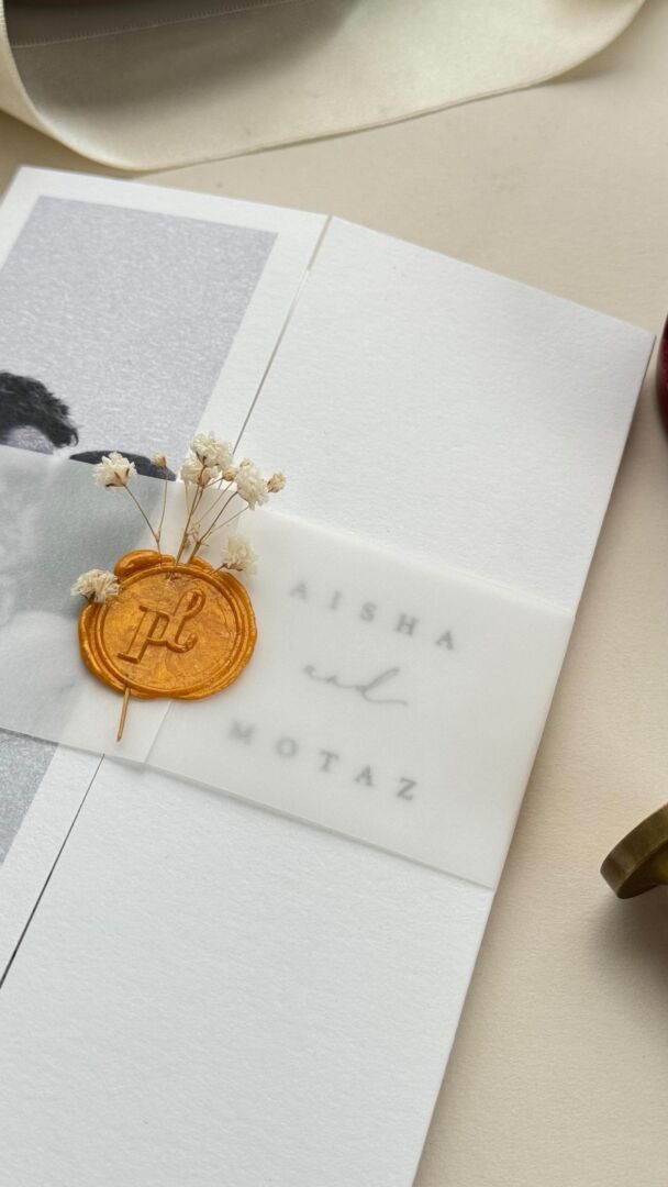 Gatefold invitation with gold wax seal and dried floral accent, held in hand.