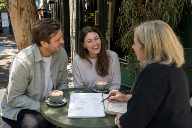 A naturally beautiful couple in their late twenties sitting across a small outdoor cafe table from their marriage celebrant in Australia.