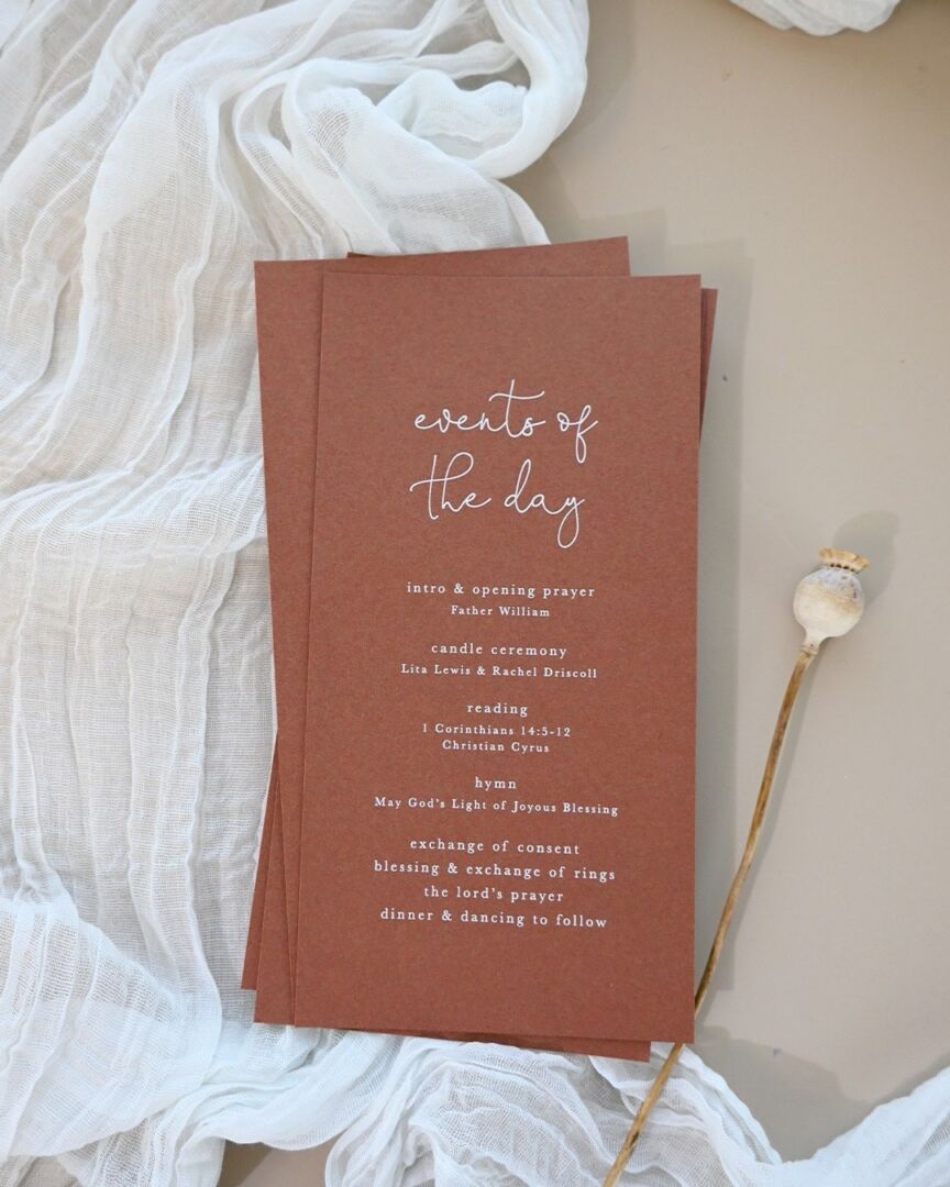 Terracotta wedding programs printed with 'events of the day' sit on white tulle with a dried flower stem.