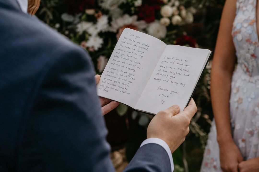 Close-up of a person's hands holding open a small vow booklet with handwritten text, ceremony setting with flowers softly blurred in background.