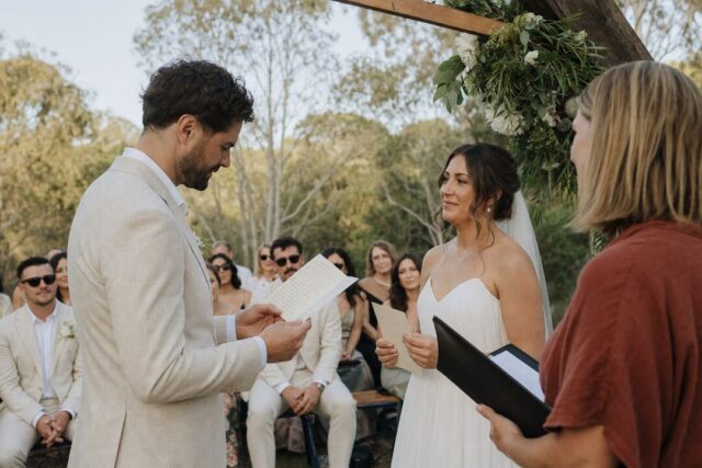A groom reading his handwritten vows from a cream vow card tilted up toward his face at an outdoor Australian ceremony.