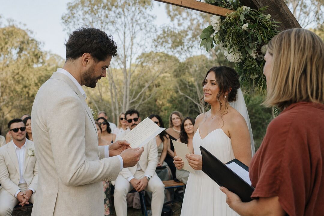 A groom reading his handwritten vows from a cream vow card tilted up toward his face at an outdoor Australian ceremony.