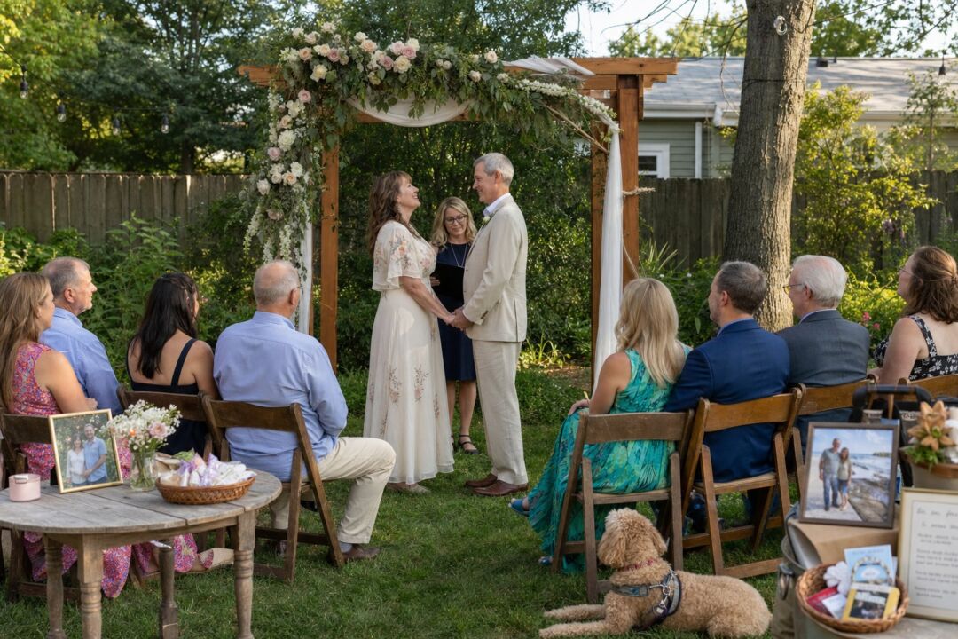 A couple in their 50s holding hands and facing each other at an intimate outdoor ceremony beneath a flower-draped arch.