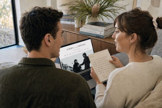 A couple on their couch reading a handwritten note beside a laptop showing their Paperlust wedding website.