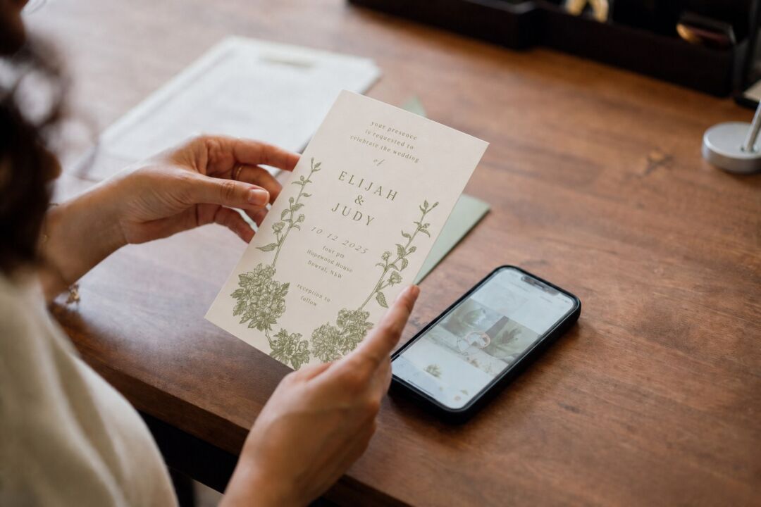 A woman holding a printed Paperlust wedding invitation beside a phone showing the couple's wedding website.