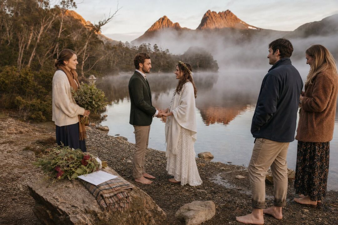 A couple standing barefoot on the edge of a misty Tasmanian mountain lake at sunrise, two witnesses visible nearby, dramatic alpine scenery.