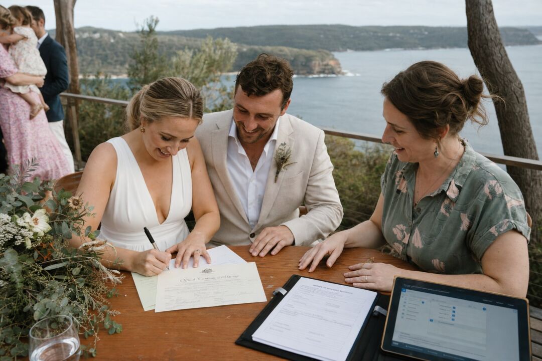 A bride in a white dress and groom in a cream suit sign marriage documents at a wooden table overlooking a scenic lake and forested landscape.