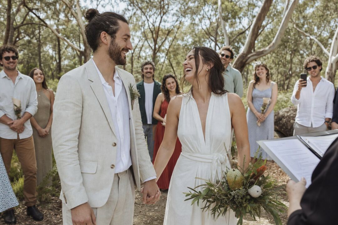 A couple in relaxed linen wedding attire holding hands and laughing during an outdoor elopement ceremony in a sun-lit eucalyptus grove.
