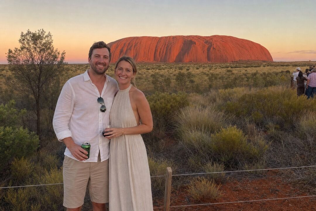 A couple standing at the base of Uluru at sunset, the monolith filling the background, desert spinifex in the foreground, warm orange light.