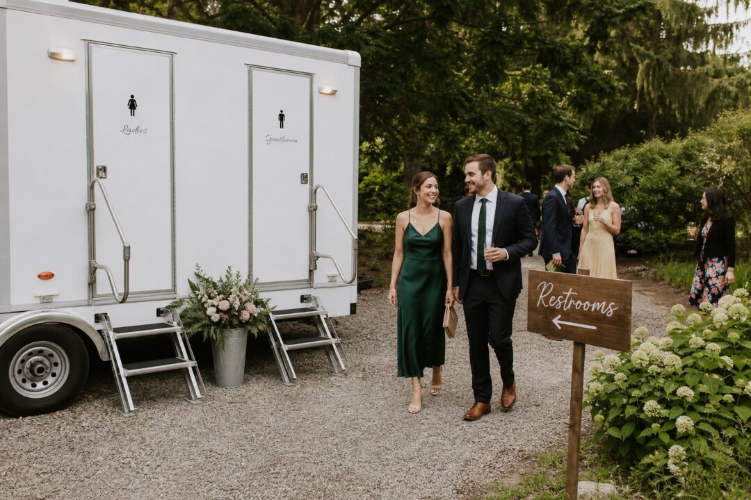 A couple walks past a white portable restroom trailer at an outdoor wedding venue surrounded by green trees and flowering shrubs.
