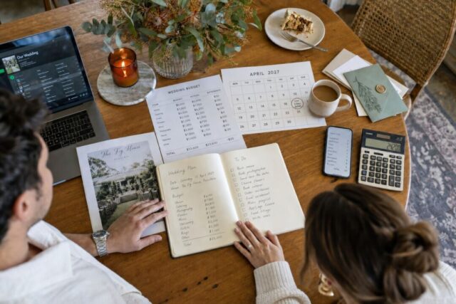A couple reviews wedding planning documents and budget spreadsheets at a wooden table with coffee, flowers, and a laptop.
