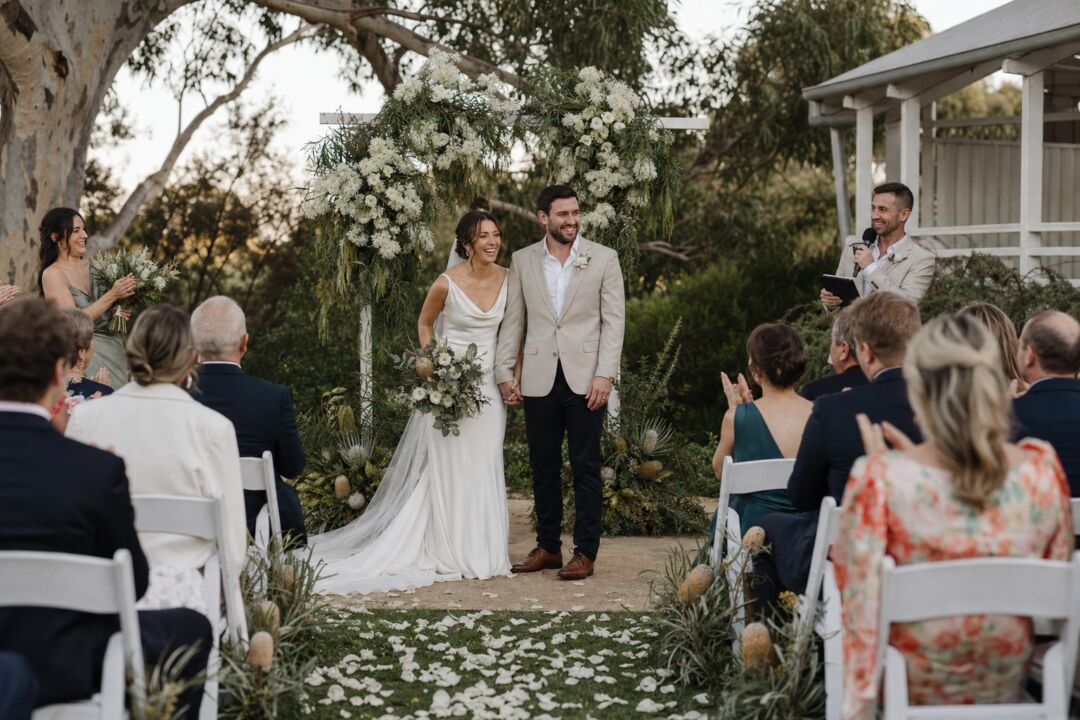 A bride and groom standing at an outdoor ceremony under a white floral arch.