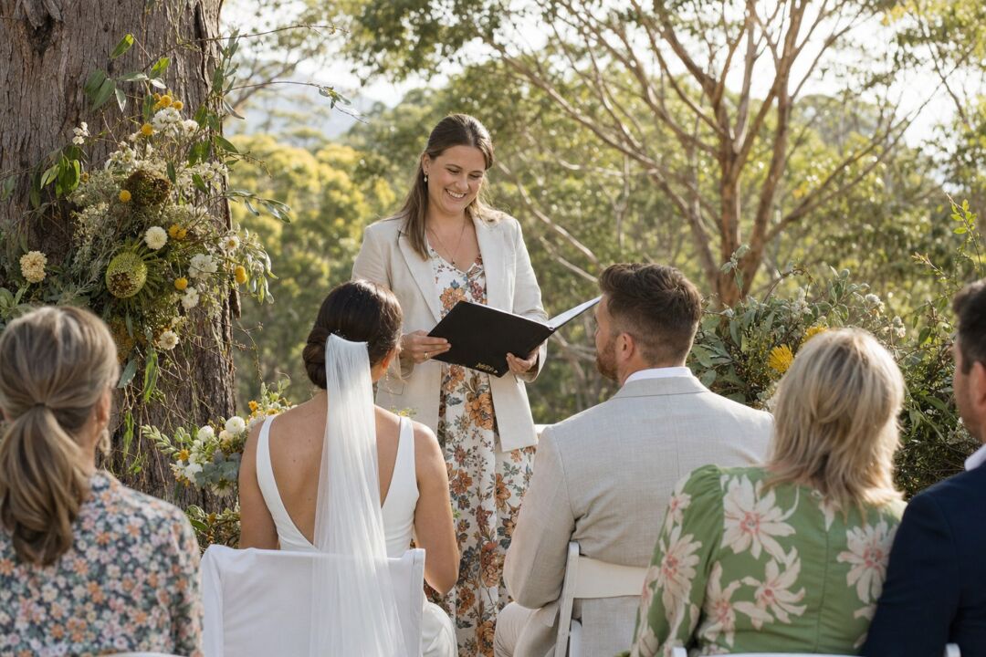A smiling marriage celebrant standing before a seated couple at an outdoor garden ceremony, holding an open ceremony folder.