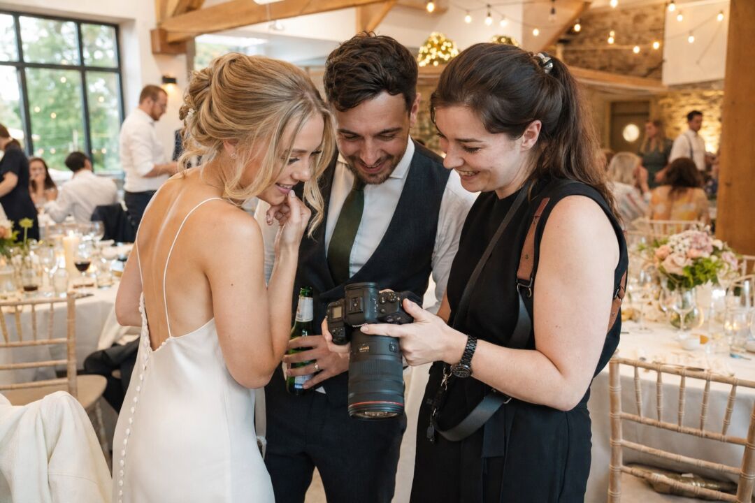 A wedding photographer reviewing images on the back of a camera at a reception venue, with warmly lit tables visible in the background.