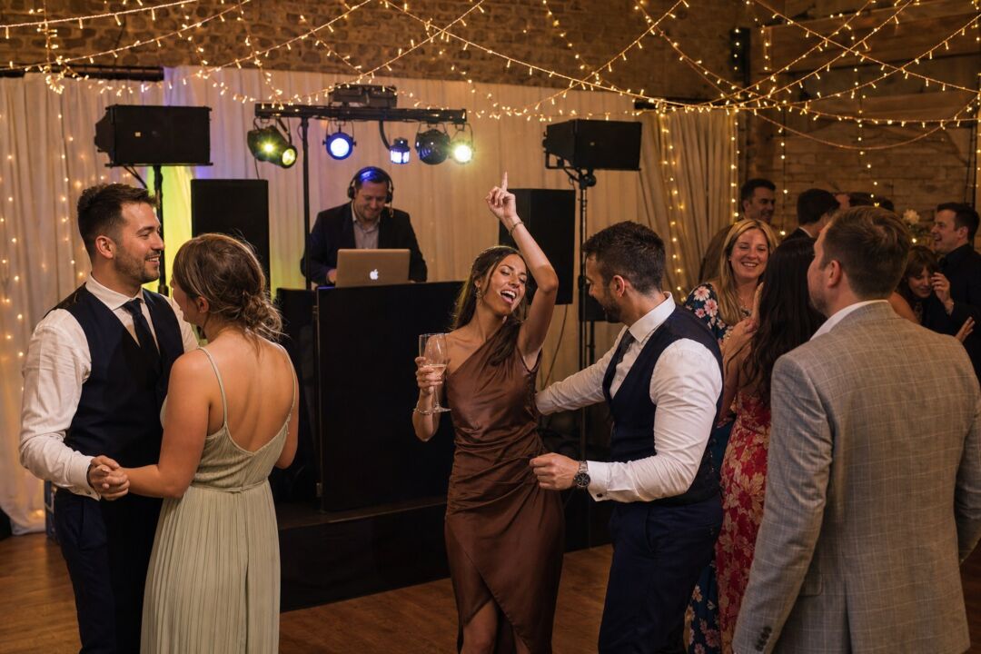 A wedding DJ booth set up inside a decorated reception hall at night, with warm string lights overhead and guests dancing in the foreground.