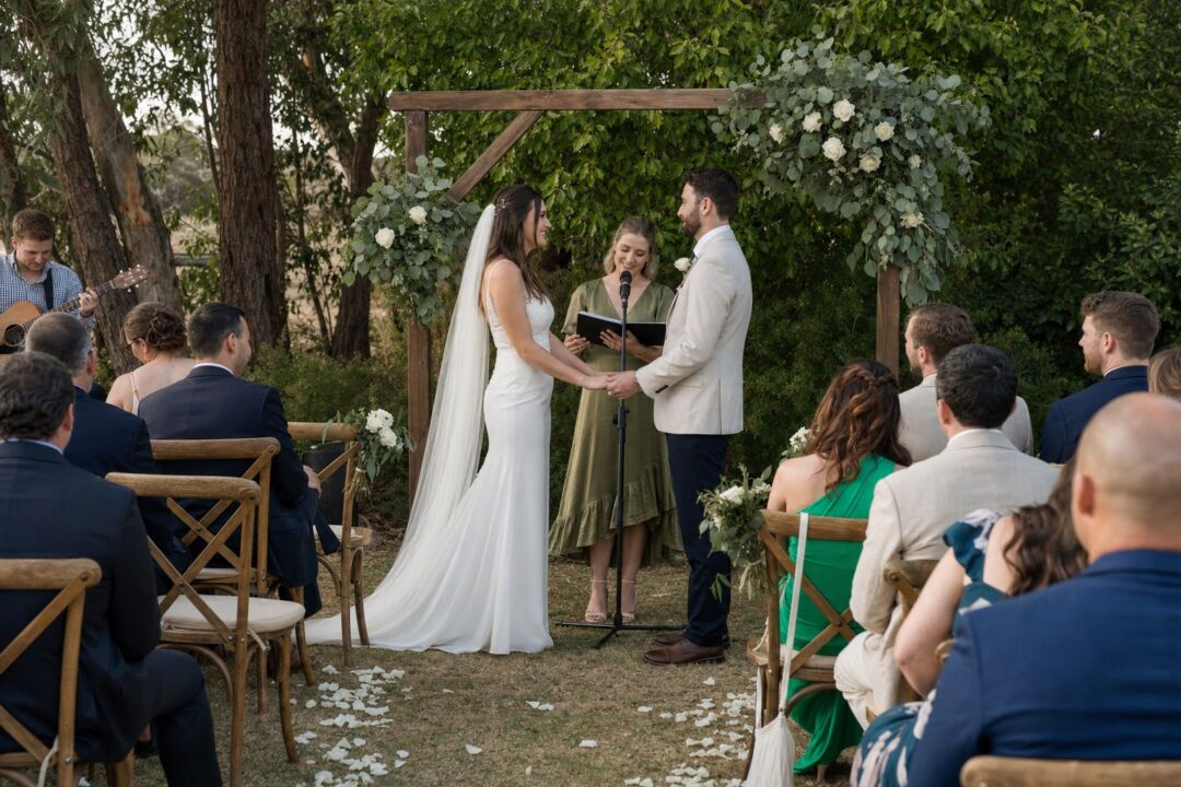 A wedding ceremony outdoors at a dry-hire venue, a standing microphone positioned at a timber arbor decorated with greenery.