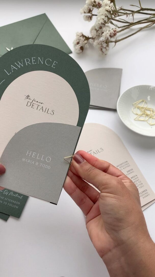 A Paperlust wedding invitation suite in sage and stone, held in one hand over a pale surface with dried florals.