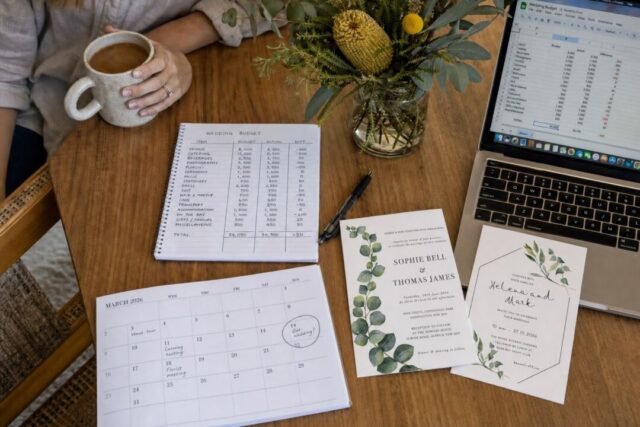 A wooden desk displays wedding planning materials including budget notes, invitations, a calendar, flowers in a vase, coffee, and a laptop.
