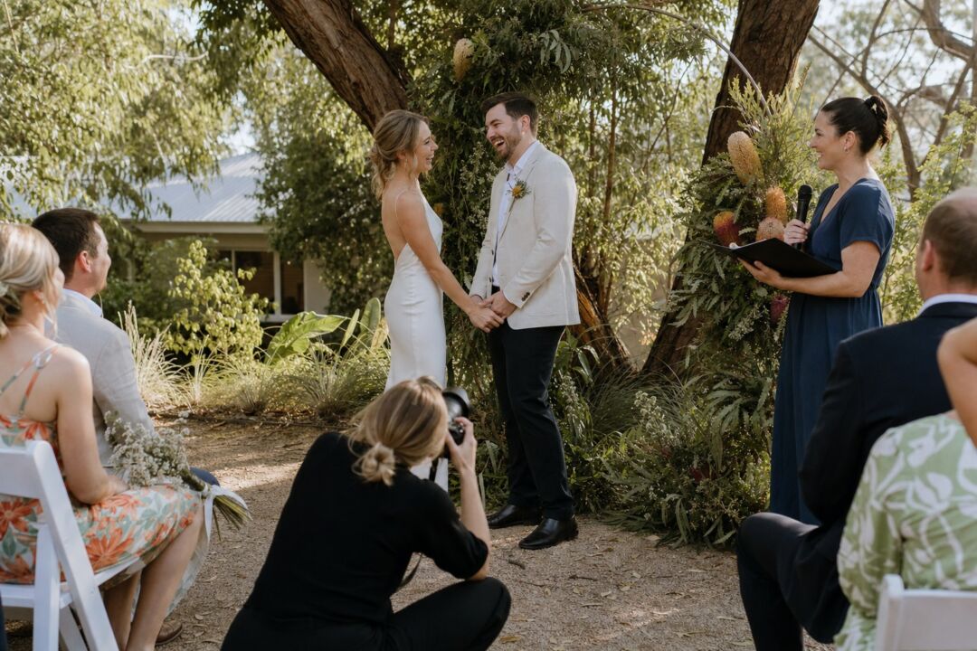 A couple exchanges vows beneath a leafy tree during an outdoor garden wedding ceremony with guests seated around them.
