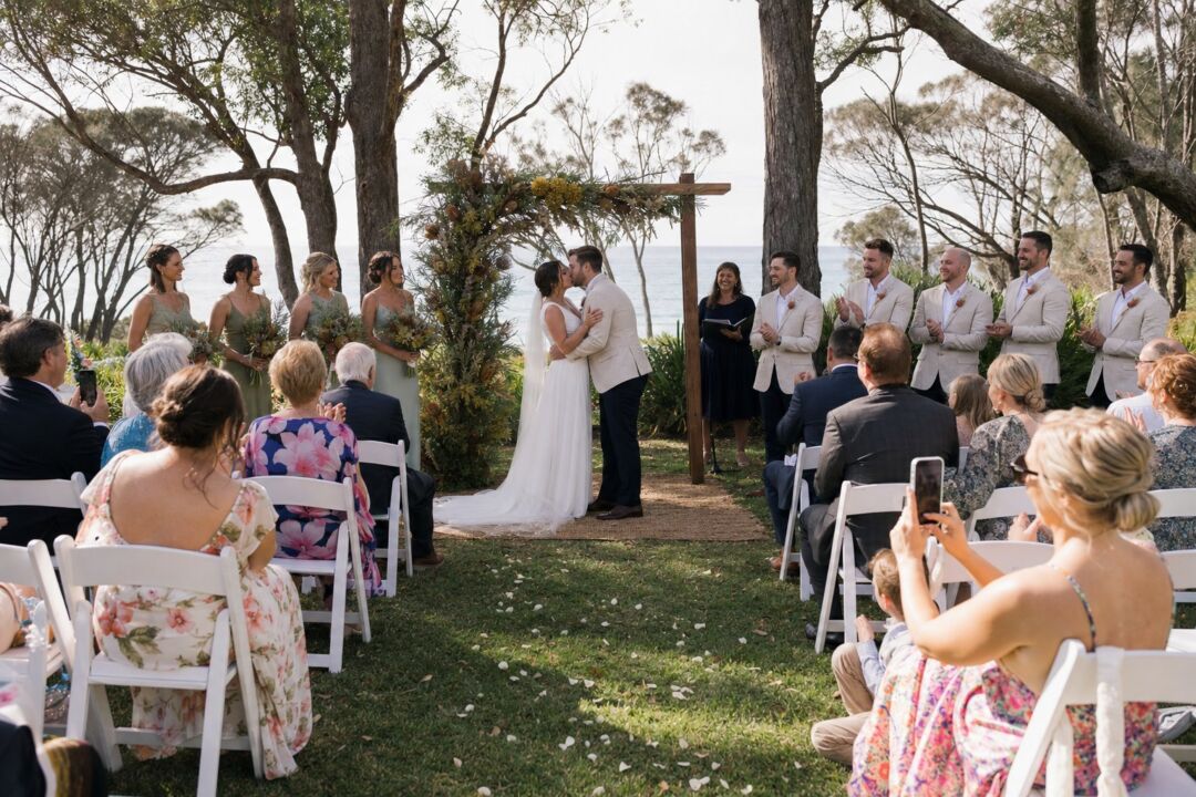 A ceremony setup in a sun-lit coastal park with white folding chairs arranged on green grass, a simple floral arch of native Australian flowers.