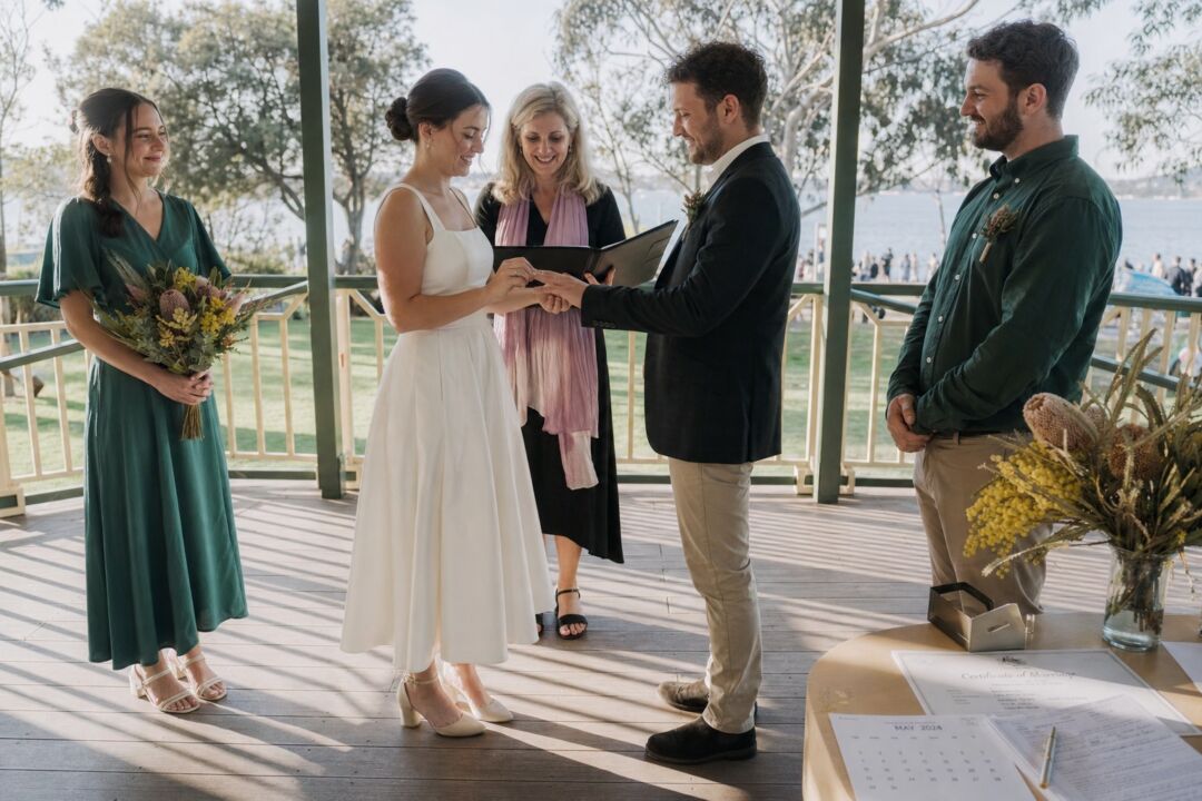 A couple and celebrant at the moment of the ring exchange in a public park rotunda, two witnesses standing beside them.