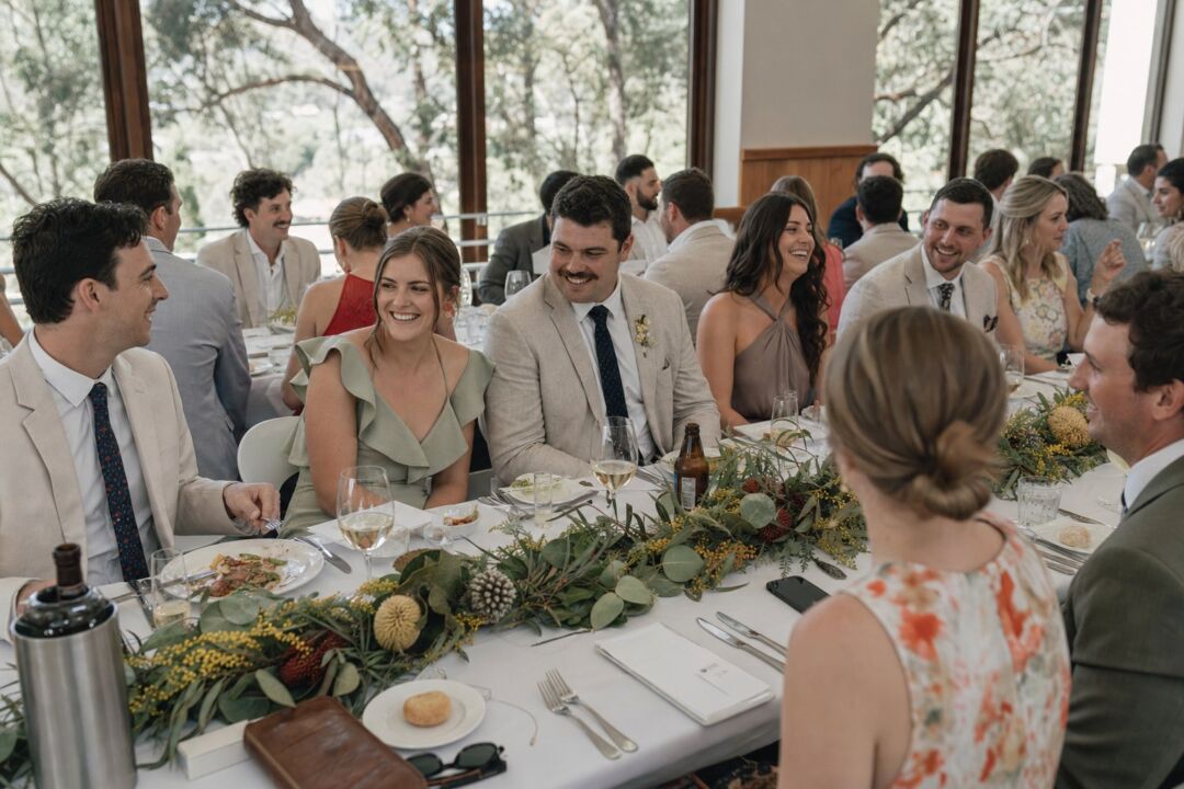 A long shared table set for a wedding lunch in a light-filled restaurant function room, native greenery as a centerpiece.