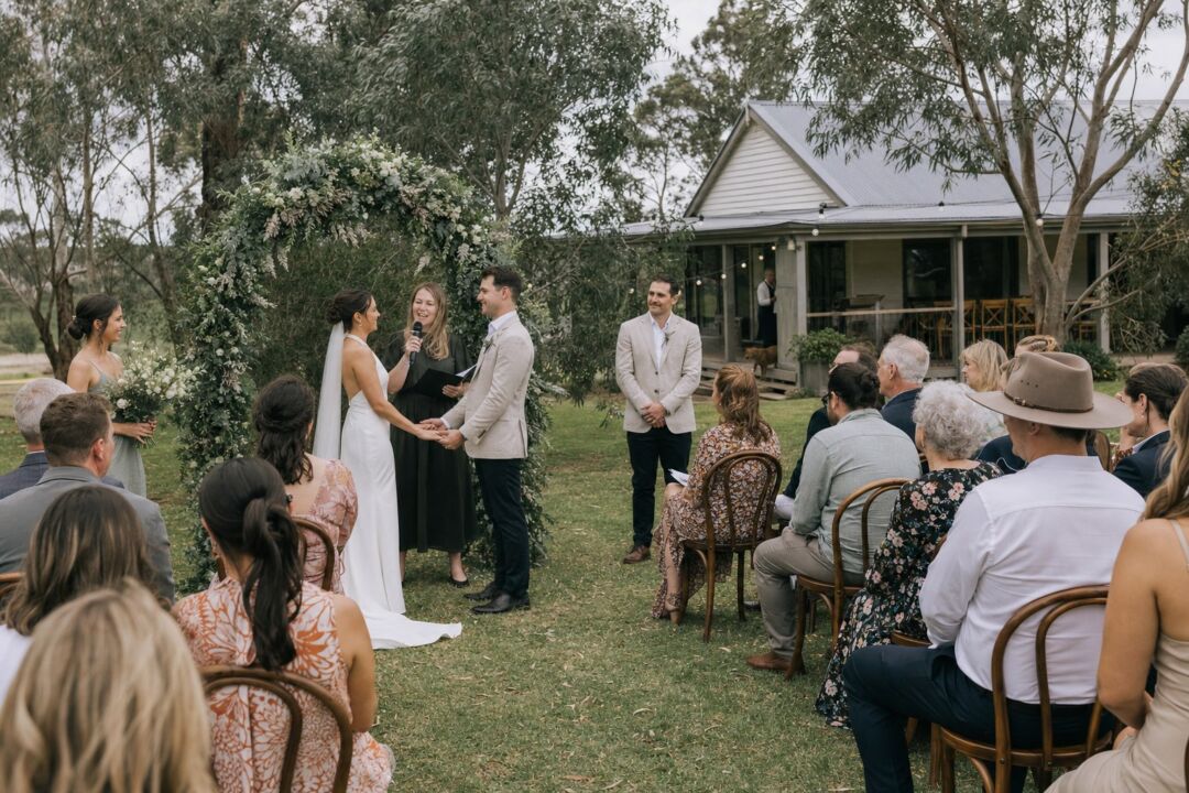 A garden ceremony on a private rural property, guests seated on timber chairs among established fruit trees.