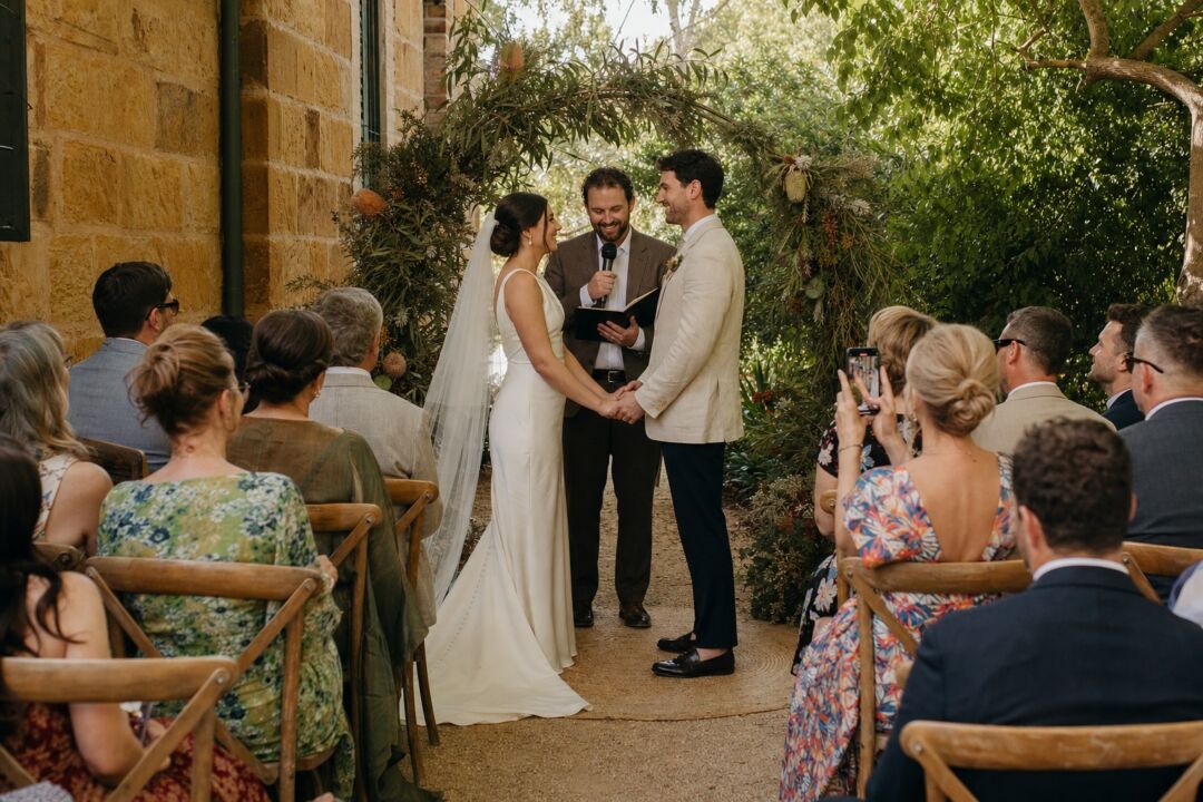 An intimate outdoor wedding ceremony at a garden estate, 28 guests on timber chairs arranged in two rows beside a sandstone wall.