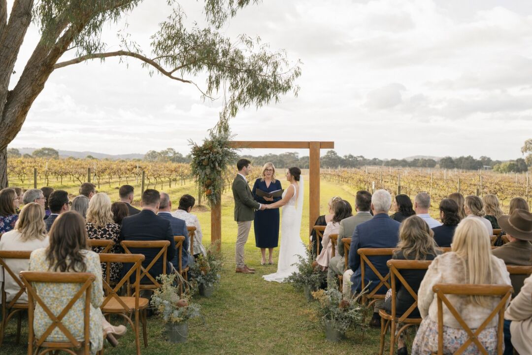A couple exchanging vows at a vineyard ceremony, 30 wooden chairs in two rows facing a simple arch.