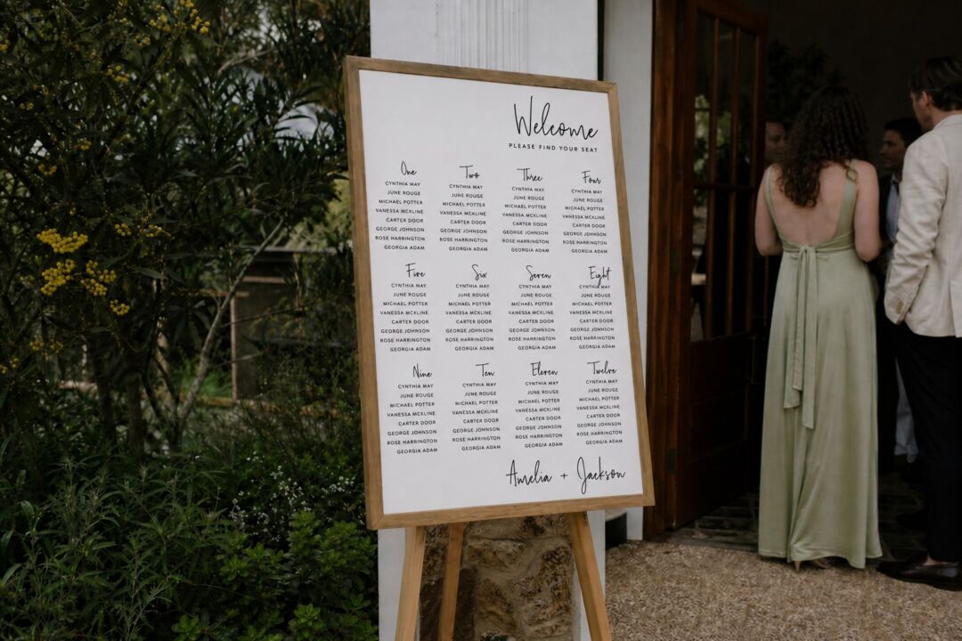 A wooden-framed seating chart displays guest names organized by table at an outdoor wedding venue surrounded by greenery.