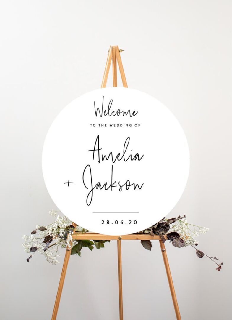 A Paperlust circular wedding welcome sign on a timber easel with dried florals at its base.