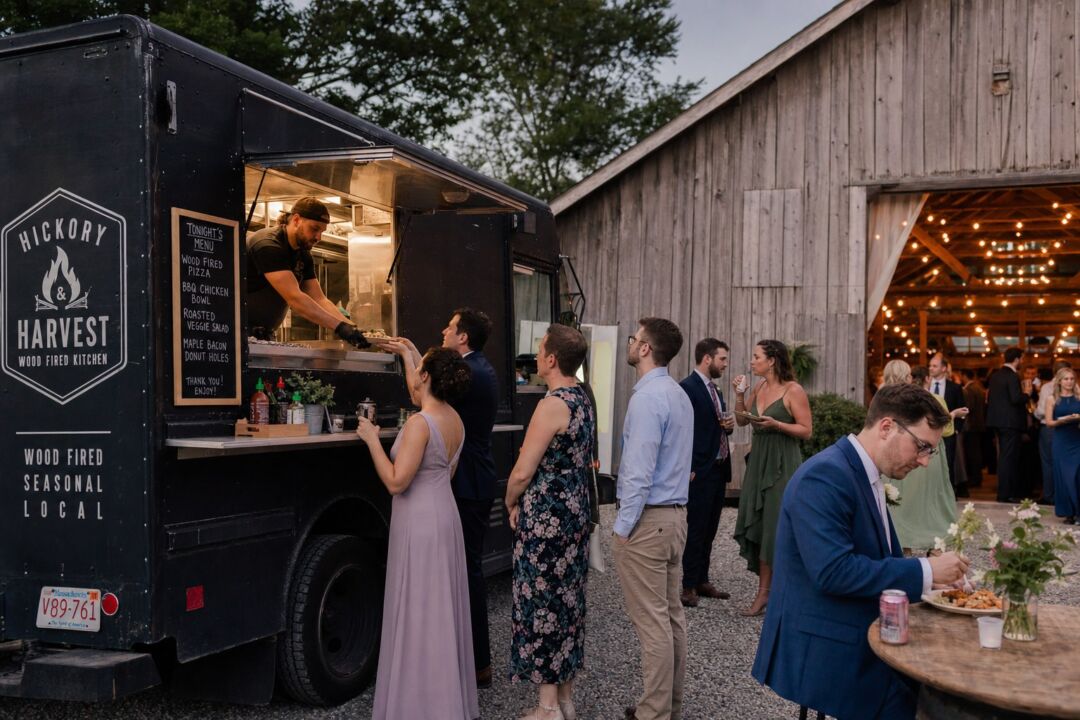 A food truck parked on a gravel area beside a timber barn at dusk, with wedding guests gathering around.
