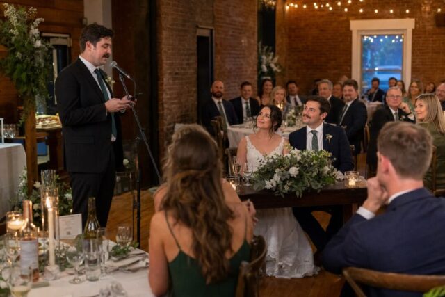 A wedding MC standing at a microphone in a warmly lit reception hall.