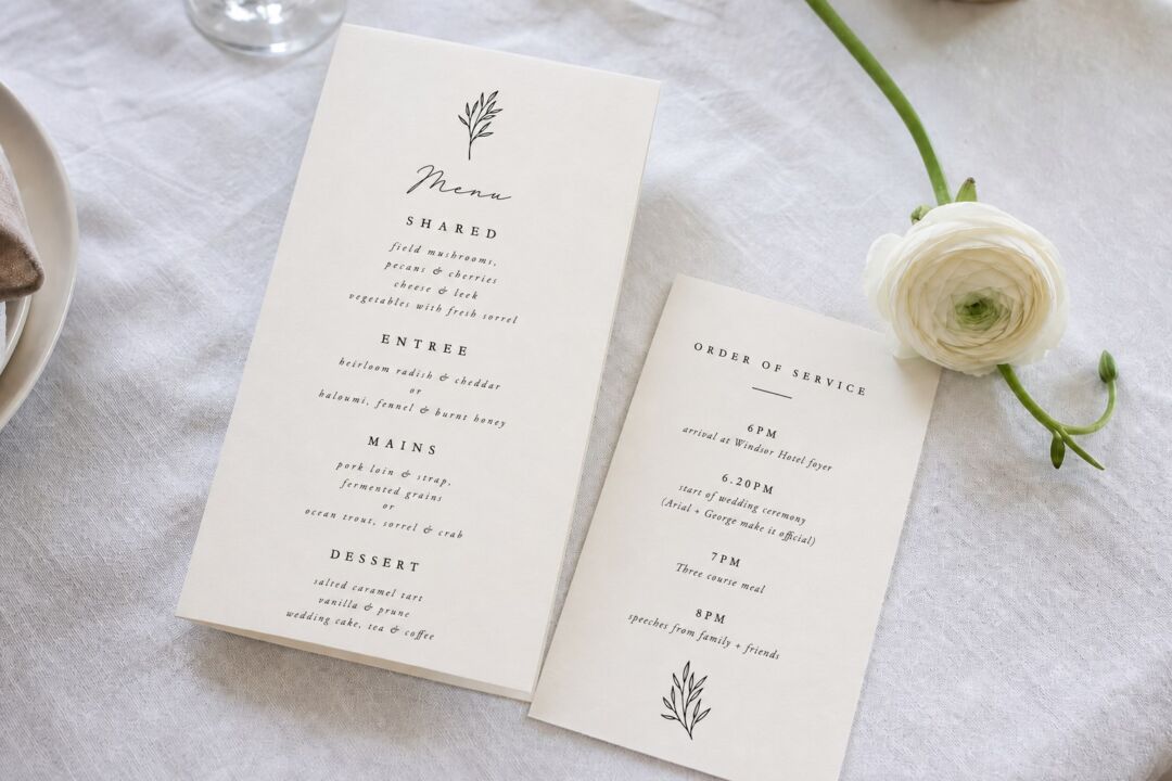 Two cream wedding menu cards with botanical illustrations sit on a white linen tablecloth beside a white ranunculus flower.