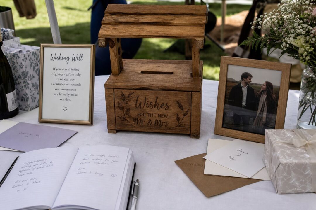 A small handmade timber wishing well box with a coin slot in the lid, placed on a white linen tablecloth at an outdoor reception.