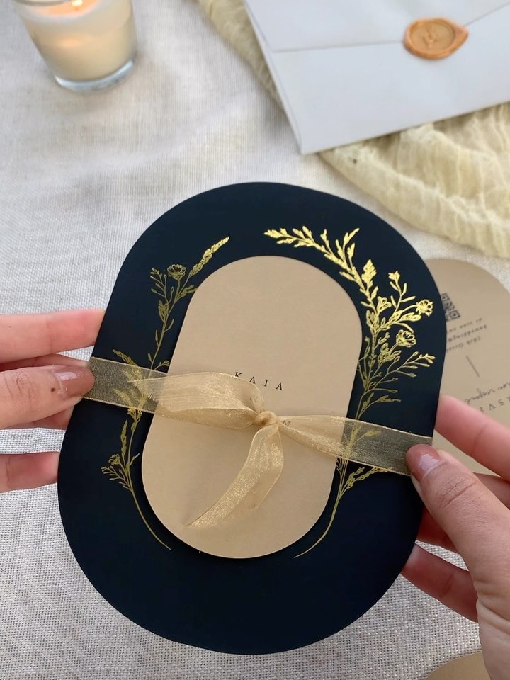 A navy and gold foil oval wedding invitation suite tied with ribbon, held in two hands beside a wax sealed envelope and a printed details card.