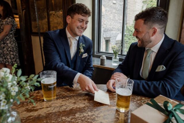 A groom sliding a handwritten card across a worn wooden pub table toward his best mate, two glasses of beer in frame, both laughing.