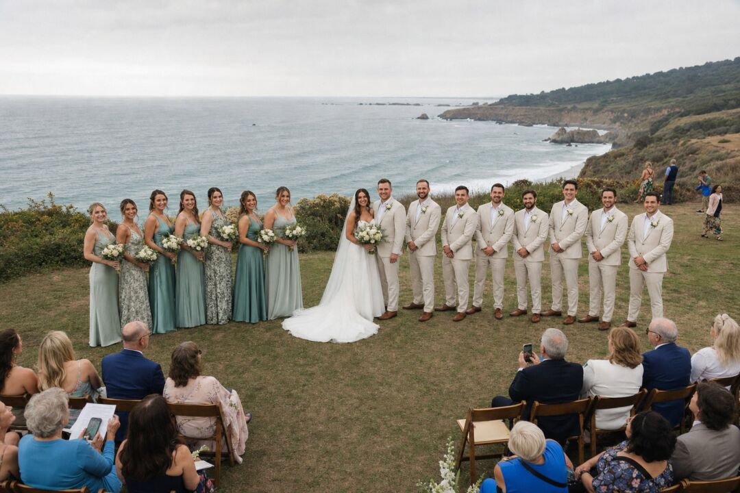 A large wedding party of eight on each side arranged in a curved arc at an outdoor clifftop ceremony with ocean views.