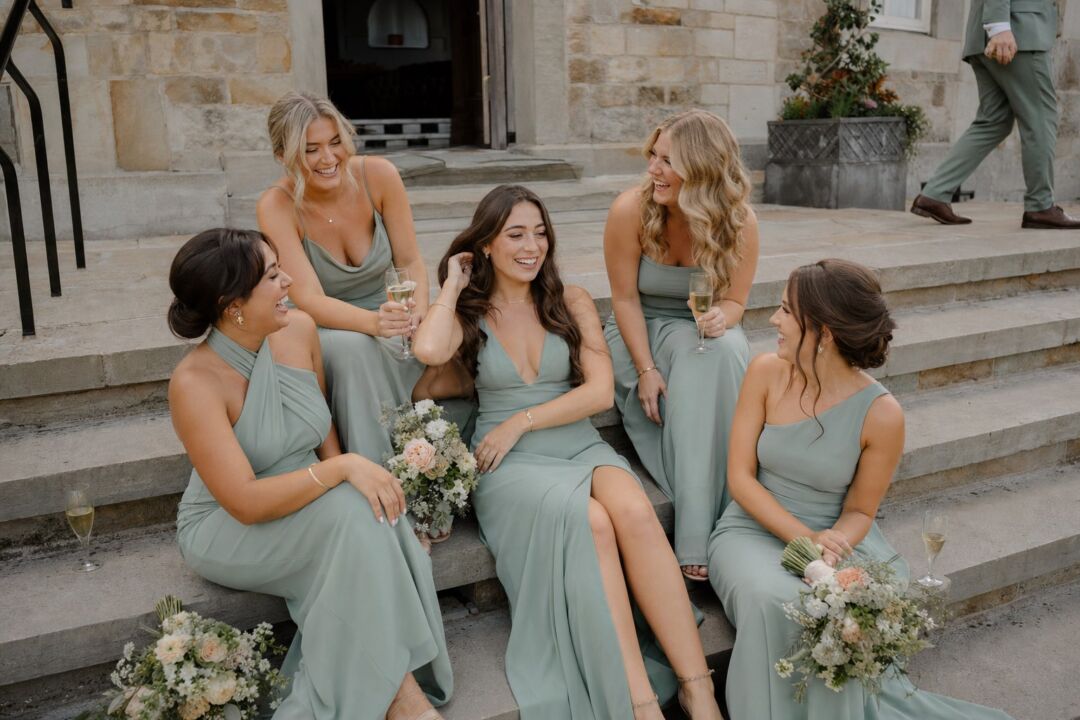 A group of five young women in coordinated sage bridesmaid dresses seated casually on wide stone venue steps, laughing together.
