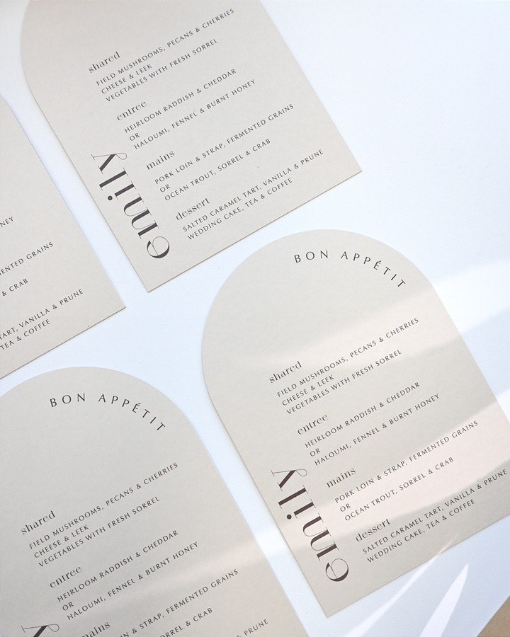 A neutral wedding stationery flat lay: arch-topped ivory menu cards on a greige surface in soft directional light.