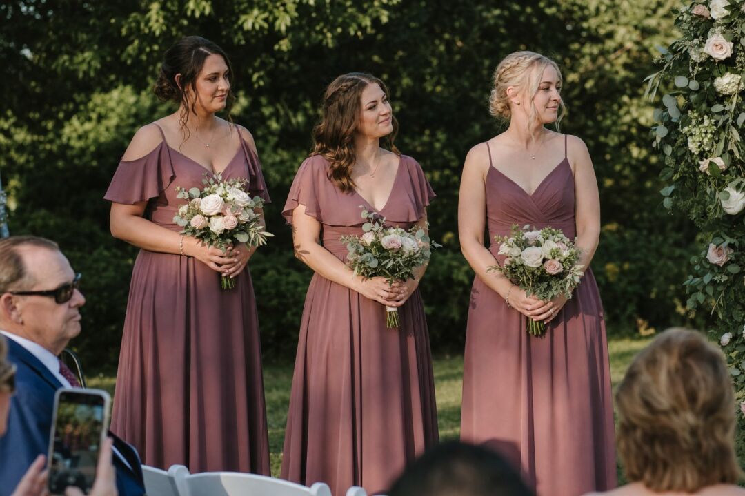An outdoor ceremony scene in warm late-afternoon light showing bridesmaids in dusty mauve dresses holding bouquets of blush, ivory.