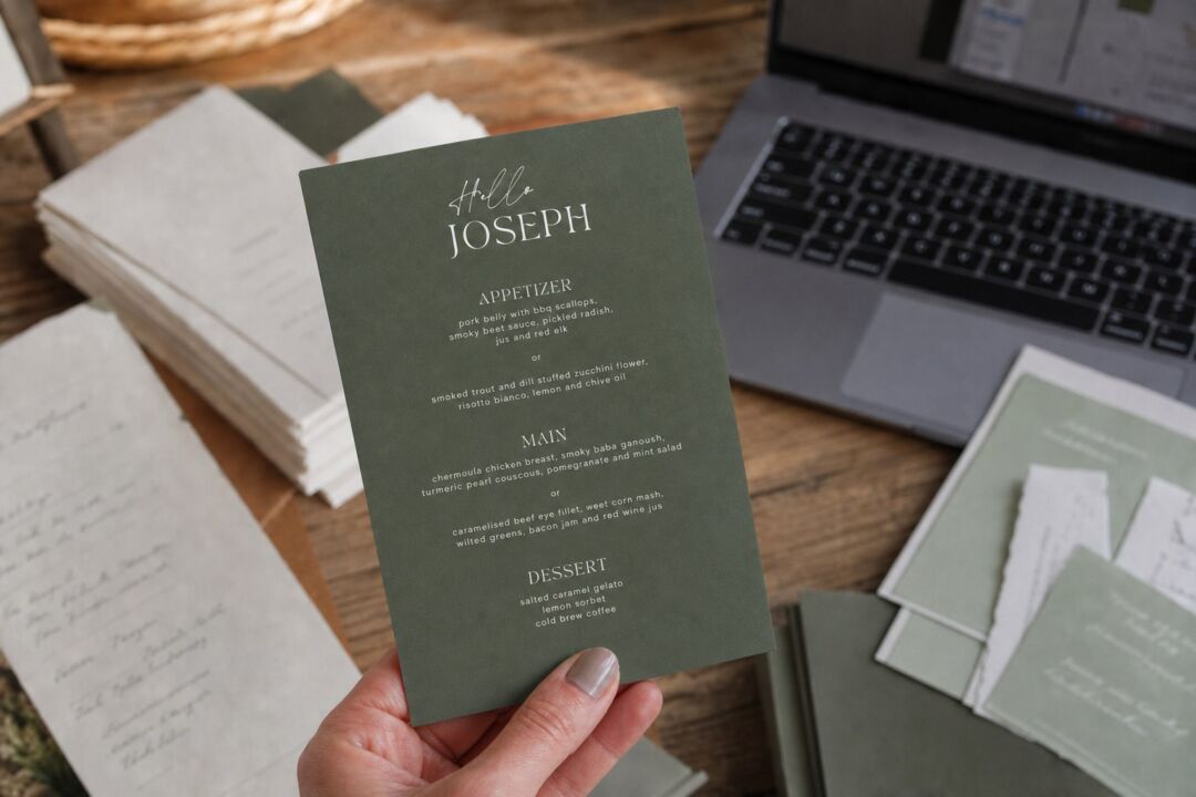 A hand holding a Paperlust sage green wedding menu printed in white ink, beside a laptop and a stack of printed paper samples.