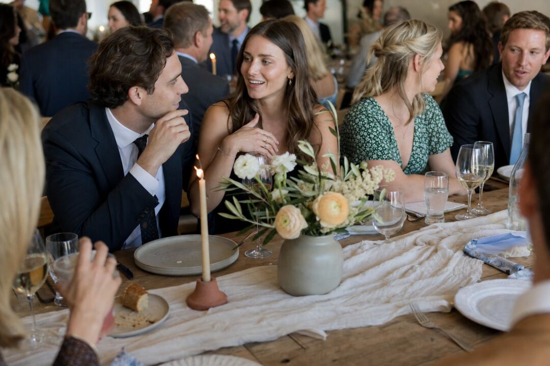 A wedding tablescape in a neutral rustic style showing a matte ceramic vessel with loose ranunculus and olive branches.