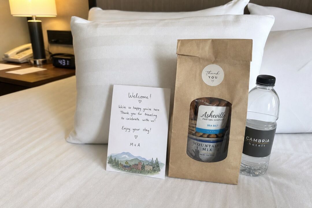 A small handwritten welcome card propped on a hotel pillow next to a kraft paper bag with local snacks.