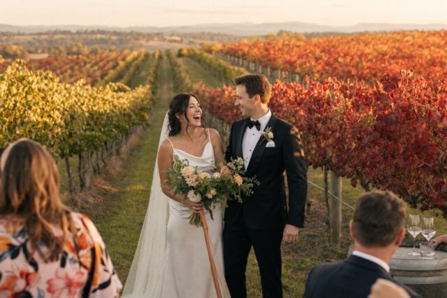 A bride and groom stand together in a vineyard during autumn, surrounded by red and golden grapevines with guests visible in the foreground.