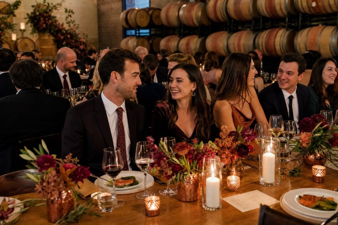 Guests in formal attire enjoy dinner at a long table decorated with burgundy and orange flowers, candles.