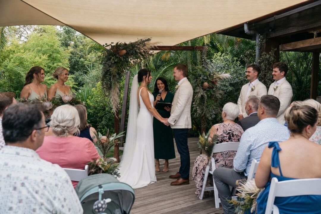 A bride and groom exchange vows under a pergola adorned with greenery and flowers, surrounded by seated guests in a lush garden setting.