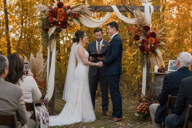 A bride and groom exchange vows under an ornate wooden arch decorated with ivory fabric, burgundy and orange flowers, and pampas grass.
