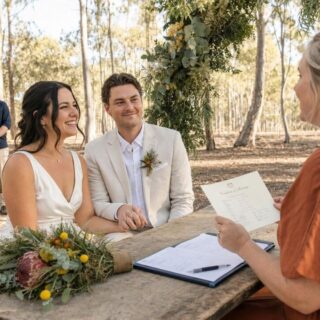 A bride and groom exchange vows with an officiant at an outdoor woodland ceremony decorated with greenery and flowers.